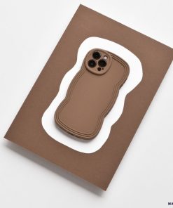 A smartphone with the NAKD Curvy Phone Case - Coffee by Coconut Lane sits on a white paper cutout, which rests atop a larger rectangular brown surface. The entire setup is on a light gray background.