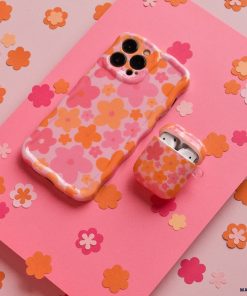 A Wavy Phone Case - Pink & Orange Bloom by Coconut Lane and an AirPods case, both covered in matching floral designs with pink, orange, and peach-colored flowers, are placed on a pink background scattered with small paper flowers in similar colors. This extra protective duo adds a charming touch to your tech essentials.