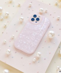 A Moonstone Pink Phone Case by Coconut Lane, featuring a pale pink hue with a pearlescent finish, is showcased on a textured pink surface. Variously sized white pearls and small multicolored beads encircle the case, creating an elegant and whimsical display.