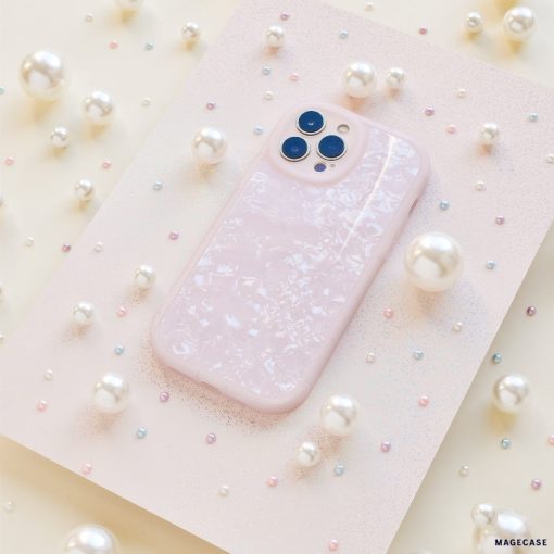 A Moonstone Pink Phone Case by Coconut Lane, featuring a pale pink hue with a pearlescent finish, is showcased on a textured pink surface. Variously sized white pearls and small multicolored beads encircle the case, creating an elegant and whimsical display.