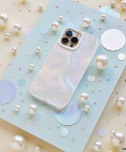 A Mother of Pearl Phone Case from Coconut Lane lies on a light blue, glittery surface surrounded by various sizes of white pearls and holographic circles. The pearlescent effect creates an aesthetic and dreamy visual effect.