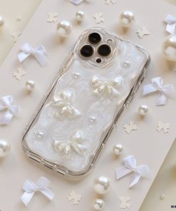 The Pearls & Bows Phone Case by Coconut Lane is adorned with white ribbon bows, pearls, and star-shaped confetti. This protective phone case showcases a pearlescent design featuring three 3D white bows and small pearl accents, all set against an elegant light-colored background.