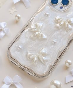 A smartphone in a Coconut Lane Pearls & Bows Phone Case, adorned with pearlescent bows and pearls, rests on a light surface scattered with white ribbons and small pearls. The phone's dual-camera lenses are visible at the top right. The image exudes a soft, elegant aesthetic.