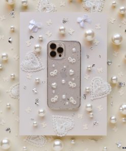 A Coquette Dreams Phone Case by Coconut Lane for the iPhone 15 lies in the center of a beige background adorned with pearls, lace hearts, and tiny bows. The intricate details of this extra protective case and the surrounding decorations exude a delicate, elegant aesthetic.