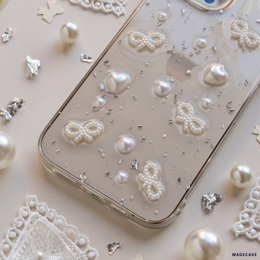 A close-up image of the Coquette Dreams Phone Case by Coconut Lane, a transparent iPhone 15 case adorned with 3D white butterfly and heart designs, as well as pearl-like embellishments. Extra protective, the phone rests on a white surface surrounded by decorative pearls, lace cutouts, and silver confetti pieces.