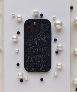 A Moonstone Black Phone Case by Coconut Lane, featuring a textured, glossy finish, is centered on a white rectangular background. The phone case is surrounded by scattered pearls in various sizes and a few black round beads, creating a contrasting and elegant arrangement that showcases its durable protection.