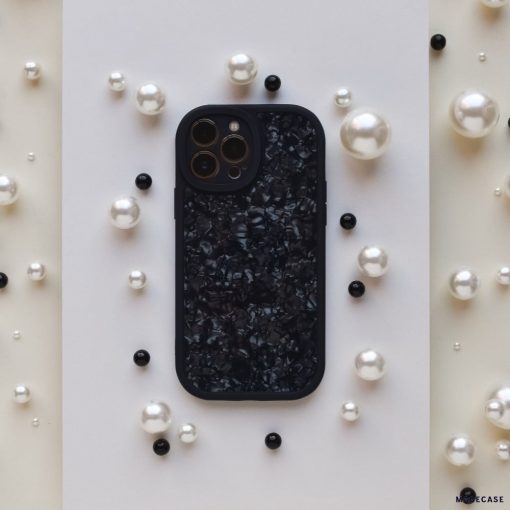 A Moonstone Black Phone Case by Coconut Lane, featuring a textured, glossy finish, is centered on a white rectangular background. The phone case is surrounded by scattered pearls in various sizes and a few black round beads, creating a contrasting and elegant arrangement that showcases its durable protection.