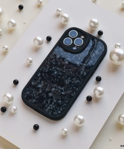 A smartphone with a dark, textured back cover lies on a white surface. Surrounding the phone are scattered white and black pearl-like beads, providing an elegant contrast to the sleek device, showcasing its sophisticated Moonstone Black Phone Case by Coconut Lane that offers durable protection.