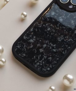A Moonstone Black Phone Case by Coconut Lane with a pearlescent pattern lies on a flat surface surrounded by scattered pearls of various sizes. The phone case has a cutout for the camera module, which is partially visible at the top right corner, offering durable protection while maintaining elegance.