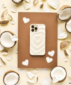 A white smartphone with the Melting Heart Phone Case - Coconut Cream by Coconut Lane lies on a brown rectangular surface. Surrounding the phone are halved coconuts and scattered coconut pieces mixed with white paper heart cutouts, all arranged on a beige background.