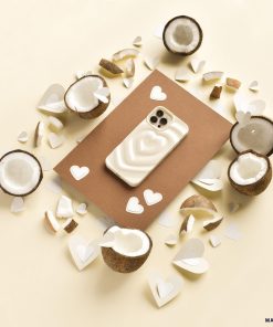 A smartphone with the Melting Heart Phone Case - Coconut Cream from Coconut Lane lies on a brown rectangular sheet. Surrounding it are halved coconuts and heart-shaped paper cutouts scattered on a cream-colored surface, creating a warm, tropical, and romantic ambiance.