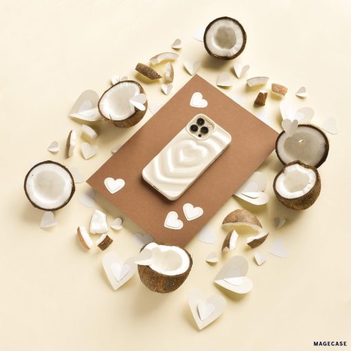 A smartphone with the Melting Heart Phone Case - Coconut Cream from Coconut Lane lies on a brown rectangular sheet. Surrounding it are halved coconuts and heart-shaped paper cutouts scattered on a cream-colored surface, creating a warm, tropical, and romantic ambiance.