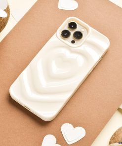 A Melting Heart Phone Case - Coconut Cream by Coconut Lane, with its smooth, pale beige case featuring a raised, 3D heart design, lies on a piece of brown paper. Surrounding the phone are small white heart-shaped cutouts and halved coconuts. The background showcases cream-colored and various brown tones.