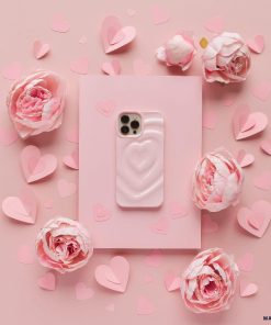 A pink smartphone adorned with Coconut Lane's Melting Heart Phone Case in Peony Pink is centered on a pink background. The phone is encircled by pink paper hearts of various sizes and clusters of light pink flowers, creating a romantic and delicate ambiance.