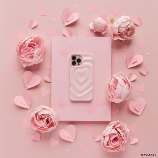 A pink smartphone adorned with Coconut Lane's Melting Heart Phone Case in Peony Pink is centered on a pink background. The phone is encircled by pink paper hearts of various sizes and clusters of light pink flowers, creating a romantic and delicate ambiance.