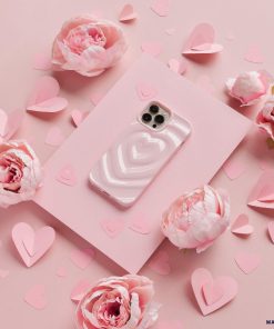A Coconut Lane iPhone adorned with a Melting Heart Phone Case in Peony Pink rests on a soft pink rectangular sheet, surrounded by delicate pink peony flowers and heart-shaped paper cutouts. The backdrop is also a gentle shade of pink, creating a romantic and cohesive aesthetic.