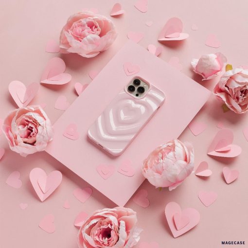 A Coconut Lane iPhone adorned with a Melting Heart Phone Case in Peony Pink rests on a soft pink rectangular sheet, surrounded by delicate pink peony flowers and heart-shaped paper cutouts. The backdrop is also a gentle shade of pink, creating a romantic and cohesive aesthetic.