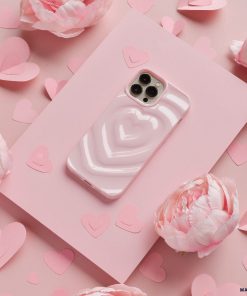 A smartphone with a Coconut Lane Melting Heart Phone Case in Peony Pink, featuring an adorable pink heart-shaped design, sits on a pink surface. It is surrounded by various sizes of pink paper hearts and delicate peony flowers. The overall theme is romantic, with a soft pink color scheme accentuated by the charming design.