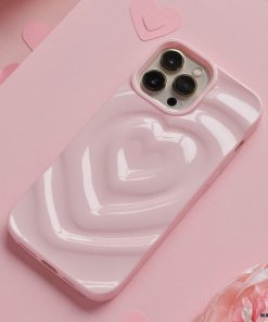 A Coconut Lane Melting Heart Phone Case in Peony Pink encases a smartphone resting on a pink surface. Multiple paper hearts are scattered around, enhancing the romantic and playful theme. The background features an array of pink shades and textures.