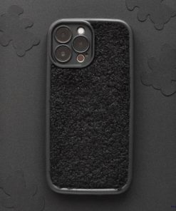 A smartphone with the Cosy Teddy Phone Case - Black from Coconut Lane is lying on a dark surface adorned with subtle floral patterns. The protective phone case features a fuzzy, fleecy texture, and the phone's camera lenses are visible in the upper left corner.