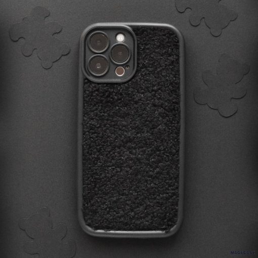 A smartphone with the Cosy Teddy Phone Case - Black from Coconut Lane is lying on a dark surface adorned with subtle floral patterns. The protective phone case features a fuzzy, fleecy texture, and the phone's camera lenses are visible in the upper left corner.
