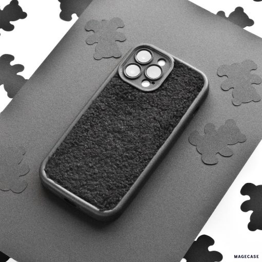A black Cosy Teddy Phone Case from Coconut Lane is displayed on a grey surface. The protective phone case has cut-outs for the camera and buttons. The background features scattered black cloud-shaped cut-outs on white paper, creating a contrasting pattern.