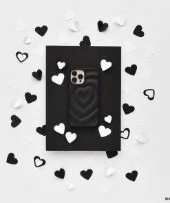 A Melting Heart Phone Case - Matte Black by Coconut Lane lies on a black rectangular backdrop. The backdrop is surrounded by scattered black and white paper hearts, creating a contrasting, monochromatic aesthetic on a white background.