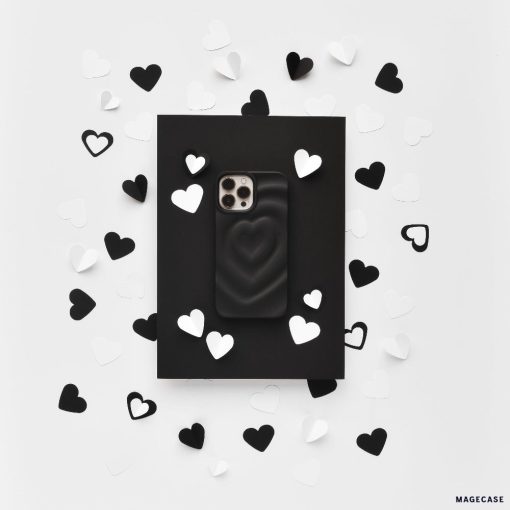 A Melting Heart Phone Case - Matte Black by Coconut Lane lies on a black rectangular backdrop. The backdrop is surrounded by scattered black and white paper hearts, creating a contrasting, monochromatic aesthetic on a white background.