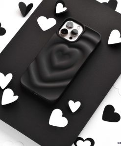 A smartphone encased in Coconut Lane's Melting Heart Phone Case - Matte Black sits on a black surface, surrounded by scattered black and white heart-shaped cutouts. The image emphasizes a monochromatic theme with a focus on the phone and the heart shapes.