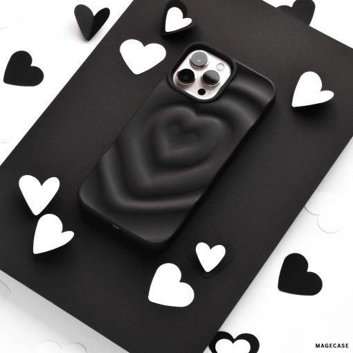 A smartphone encased in Coconut Lane's Melting Heart Phone Case - Matte Black sits on a black surface, surrounded by scattered black and white heart-shaped cutouts. The image emphasizes a monochromatic theme with a focus on the phone and the heart shapes.