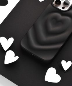 A black smartphone with the Coconut Lane Melting Heart Phone Case in Matte Black, featuring a raised heart design, is placed among scattered white and black heart-shaped cutouts on a flat surface. The phone's dual camera lenses are visible at the top.