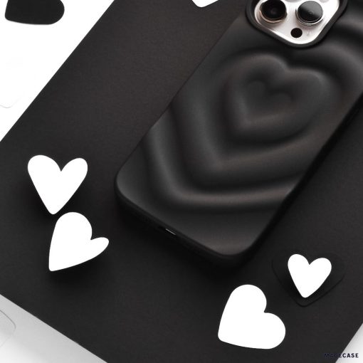 A black smartphone with the Coconut Lane Melting Heart Phone Case in Matte Black, featuring a raised heart design, is placed among scattered white and black heart-shaped cutouts on a flat surface. The phone's dual camera lenses are visible at the top.