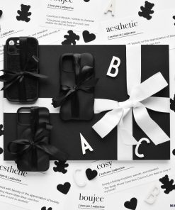A black and white display of gift-wrapped items, potentially including protective 3D Boujee Phone Cases in a sleek matte black finish from Coconut Lane, adorned with bows. The arrangement is surrounded by decorative cutouts of hearts, letters, and teddy bears. The background features printed definitions like 
