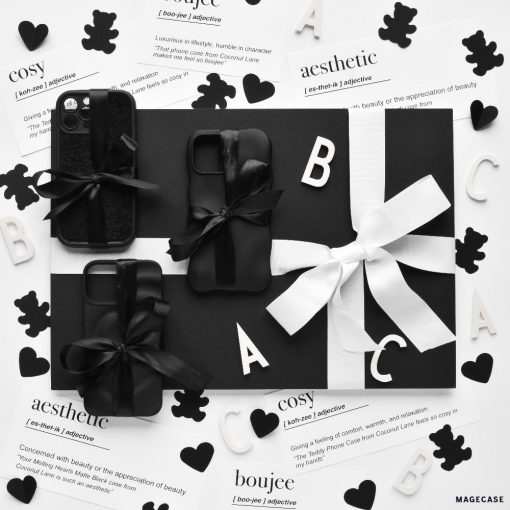 A black and white display of gift-wrapped items, potentially including protective 3D Boujee Phone Cases in a sleek matte black finish from Coconut Lane, adorned with bows. The arrangement is surrounded by decorative cutouts of hearts, letters, and teddy bears. The background features printed definitions like "cosy" and "aesthetic.