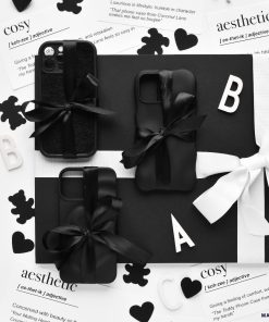 A black-and-white flat lay showcasing four Melting Heart Phone Cases - Matte Black by Coconut Lane, wrapped with black ribbons on a black background. Scattered around are cut-out letters, black paper hearts, and printed definitions of 