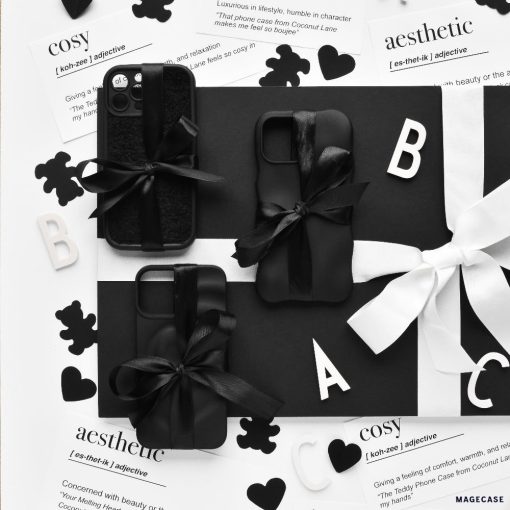 A black-and-white flat lay showcasing four Melting Heart Phone Cases - Matte Black by Coconut Lane, wrapped with black ribbons on a black background. Scattered around are cut-out letters, black paper hearts, and printed definitions of "aesthetic" and "cozy." A white ribbon bow is visible at the edge, enhancing the overall chic vibe.