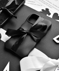 A flatlay image featuring neatly wrapped black gift boxes adorned with black and white ribbons. The central focus is on Coconut Lane's 3D Boujee Phone Case - Matte Black, elegantly tied with a black ribbon. The background showcases black and white text along with decorative elements in monochrome tones, all boasting a matte black finish.