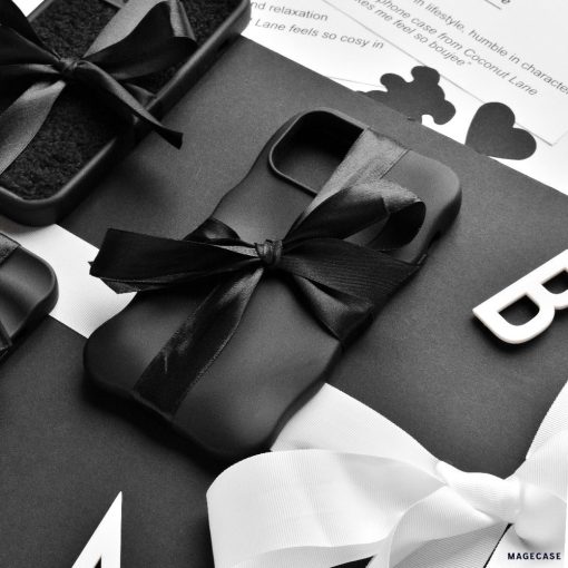 A flatlay image featuring neatly wrapped black gift boxes adorned with black and white ribbons. The central focus is on Coconut Lane's 3D Boujee Phone Case - Matte Black, elegantly tied with a black ribbon. The background showcases black and white text along with decorative elements in monochrome tones, all boasting a matte black finish.