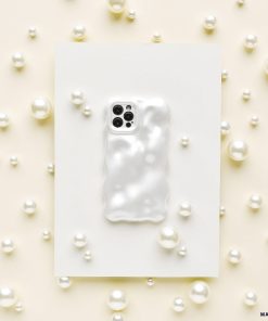 A Coconut Lane 3D Pearl Phone Case in white, featuring a glossy, wavy, pearl effect, is placed on a white rectangular sheet. The sheet is surrounded by various sizes of white pearls scattered on a light beige surface, creating an elegant and sophisticated display.