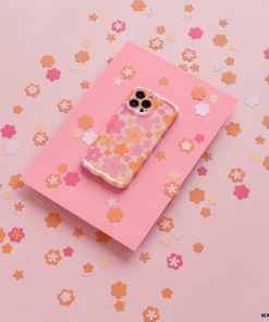 Coconut Lane's Wavy Phone Case - Pink & Orange Bloom, adorned with an extra protective pink and orange floral design, lies on a slightly darker pink sheet surrounded by small, scattered paper flowers in shades of pink, peach, and orange on a light pink background.