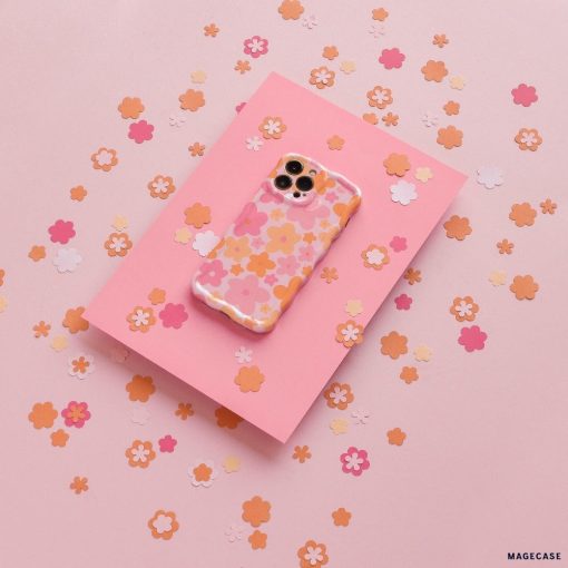 Coconut Lane's Wavy Phone Case - Pink & Orange Bloom, adorned with an extra protective pink and orange floral design, lies on a slightly darker pink sheet surrounded by small, scattered paper flowers in shades of pink, peach, and orange on a light pink background.