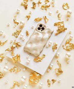 An iPhone15 adorned with Coconut Lane's 3D Pearl Phone Case - Gold rests on a white rectangular card. Scattered around are elegant gold foil pieces and 3D pearls against a beige background, crafting an extraordinarily luxurious composition.