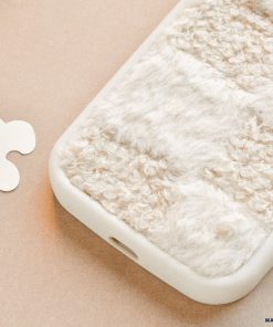 A close-up image of a Cosy Teddy Phone Case - Vanilla Biscuit from Coconut Lane, showcasing its soft, textured beige and white fur-like fabric covering. The case edges are smooth and white, providing a pleasing contrast to the cosy grip surface. The background features light, neutral tones and abstract shapes.