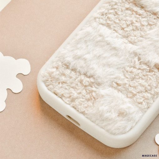 A close-up image of a Cosy Teddy Phone Case - Vanilla Biscuit from Coconut Lane, showcasing its soft, textured beige and white fur-like fabric covering. The case edges are smooth and white, providing a pleasing contrast to the cosy grip surface. The background features light, neutral tones and abstract shapes.