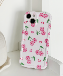 The Wavy Phone Case - Disco Cherries by Coconut Lane, featuring pink checkered cherries and small hearts on a white background, is propped up against a white surface. The glossy finish adds a touch of elegance, while in the background, part of a vase on a plate is visible.