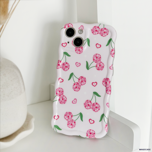The Wavy Phone Case - Disco Cherries by Coconut Lane, featuring pink checkered cherries and small hearts on a white background, is propped up against a white surface. The glossy finish adds a touch of elegance, while in the background, part of a vase on a plate is visible.