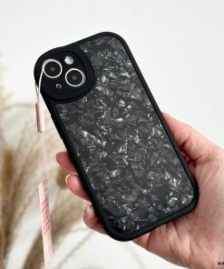 A hand holding a Moonstone Black Phone Case by Coconut Lane, featuring a textured, marble-like pattern with a small pink tag attached to it. The black phone case offers durable protection. In the background, there are blurred light-colored pampas grass.