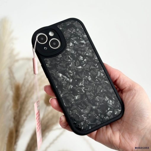 A hand holding a Moonstone Black Phone Case by Coconut Lane, featuring a textured, marble-like pattern with a small pink tag attached to it. The black phone case offers durable protection. In the background, there are blurred light-colored pampas grass.