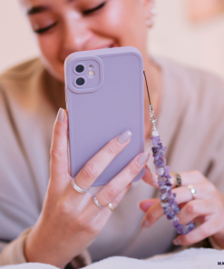 A smiling person holds a **NAKD Phone Case - Lilac** by **Coconut Lane**, complete with a beaded phone charm, emphasizing full camera protection. Their nails are manicured, and they have several rings adorning their fingers. The softly blurred background hints at a cozy indoor setting.