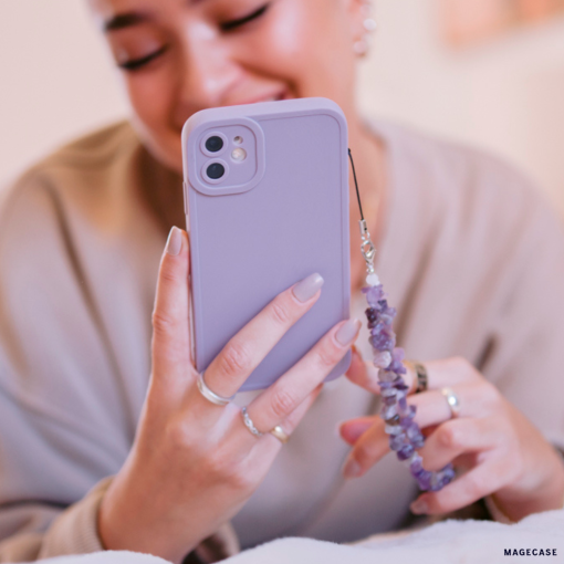 A smiling person holds a **NAKD Phone Case - Lilac** by **Coconut Lane**, complete with a beaded phone charm, emphasizing full camera protection. Their nails are manicured, and they have several rings adorning their fingers. The softly blurred background hints at a cozy indoor setting.